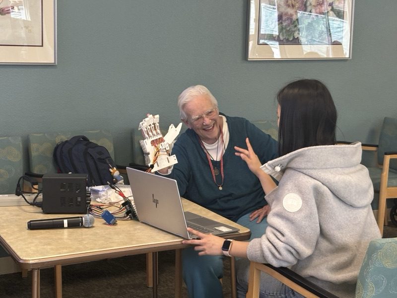 A veteran laughs while holding the Ripple exoskeleton up, talking with Yanzi across a table